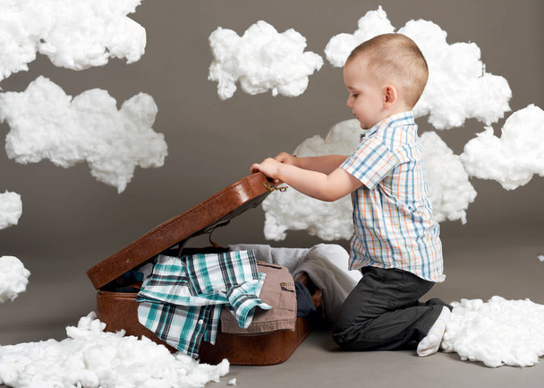 the boy is sitting in a suitcase and going on a journey, clouds of cotton wool as a decoration, gray background - Zdjęcie, obraz