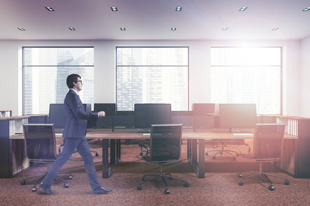 Молодой бизнесмен, прогуливающийся в открытом космосе офиса современной компании с белыми стенами, панорамными окнами, коричневым ковром и рядами деревянных компьютерных столов. 3D рендеринг макет тонированного изображения
 - Фото, изображение