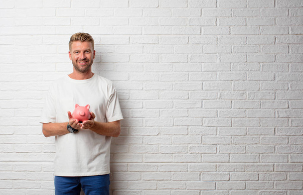 handsome blonde man with a piggy bank over a grunge wall - Photo, Image