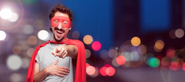 Young bearded super hero laughing hard at something hilarious and pointing towards you while holding belly with the other hand, having a good time with funny jokes. - 写真・画像