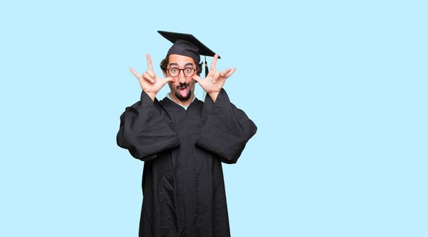 young graduated man joking, sticking tongue out with a funny, silly and playful expression, looking like a fool. - Foto, imagen