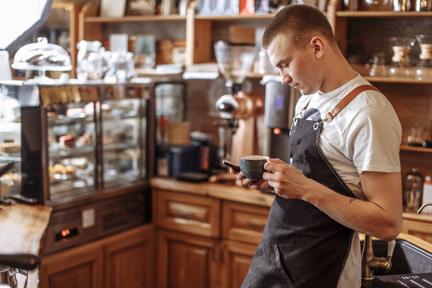 a handsome bartender drinking coffee and texting message in the smart phone - Foto, Imagen