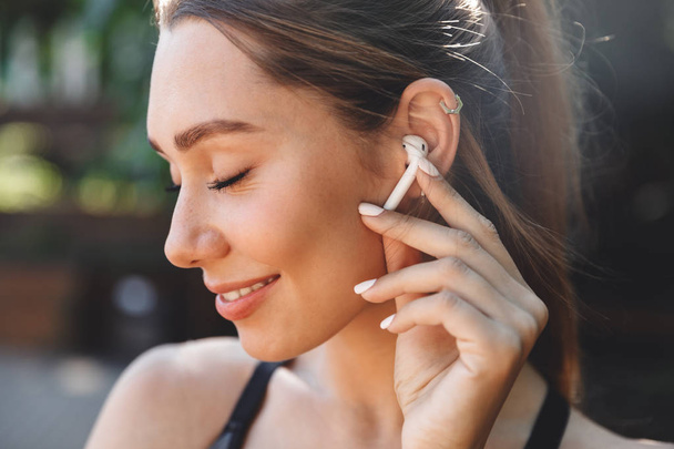 Close up portrait of a delighted young fitness girl listening to music through wireless earphones outdoors - Photo, image