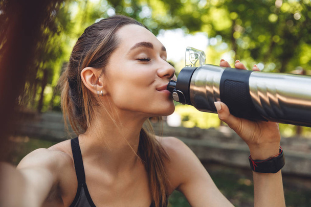 Portrait of a pretty young fitness girl taking a selfie while sitting outdoors and drinking water from a bottle - Фото, изображение