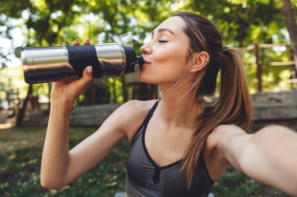 Portrait of a lovely young fitness girl taking a selfie while sitting outdoors and drinking water from a bottle - Фото, изображение