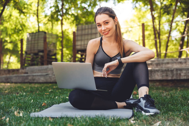 Portrét usmívající se dívky mladé fitness pomocí přenosného počítače v sedle na rohoži venku - Fotografie, Obrázek