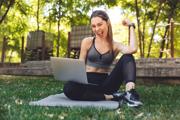 Portrait of a smiling young fitness girl using laptop computer while sitting on a mat outdoors and flexing biceps - Foto, afbeelding