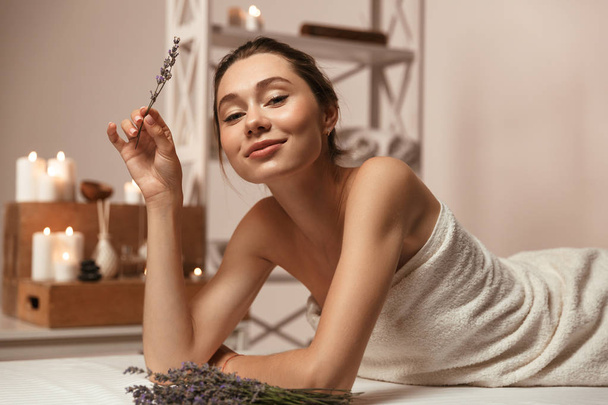 Image of amazing young beautiful woman lies in bathroom over candles holding lavender. - Foto, imagen