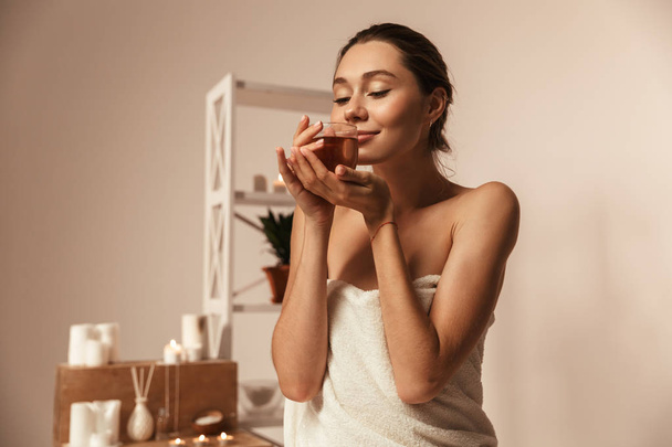 Portrait of a delighted young woman wrapped in a towel drinking herbal tea in a glass at the spa center - Foto, afbeelding