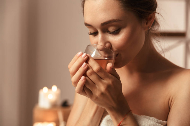 Close up portrait of an attractive young woman wrapped in a towel holding herbal tea in a glass at the spa center - Foto, Imagem