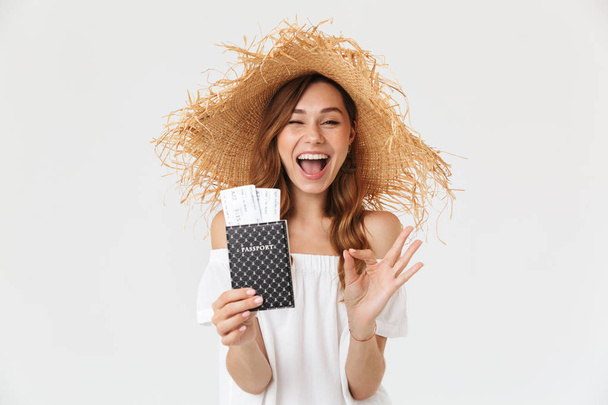 Portrait of excited woman 20s wearing big straw hat rejoicing while holding passport with tickets isolated over white background - Foto, imagen