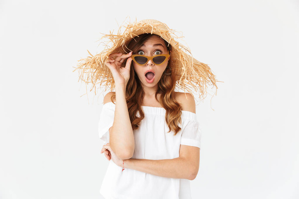Portrait of surprised pretty woman 20s wearing big straw hat looking at camera from under sunglasses isolated over white background - Foto, immagini