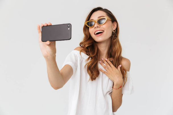 Photo of cheerful stylish woman 20s wearing sunglasses and jewelry taking selfie on smartphone isolated over white background - Photo, image