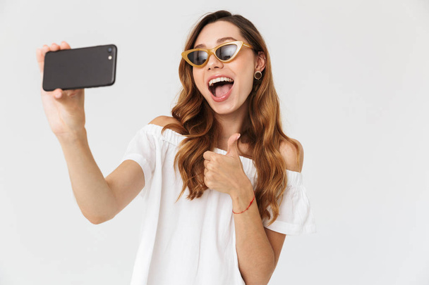 Photo of cheerful stylish woman 20s wearing sunglasses and jewelry taking selfie on smartphone and showing thumb up isolated over white background - Foto, Imagem