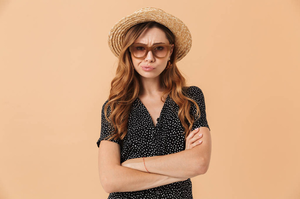 Portrait of disappointed woman 20s wearing straw hat and sunglasses standing with arms crossed isolated over beige background - Zdjęcie, obraz