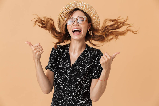 Portrait of joyful pretty woman 20s wearing straw hat and sunglasses laughing with shaking hair isolated over beige background - Foto, imagen