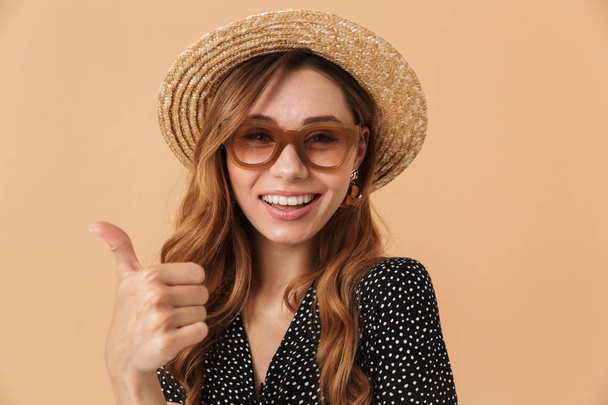 Portrait of lovely woman 20s wearing straw hat and sunglasses smiling and showing thumb up isolated over beige background - Foto, Imagen