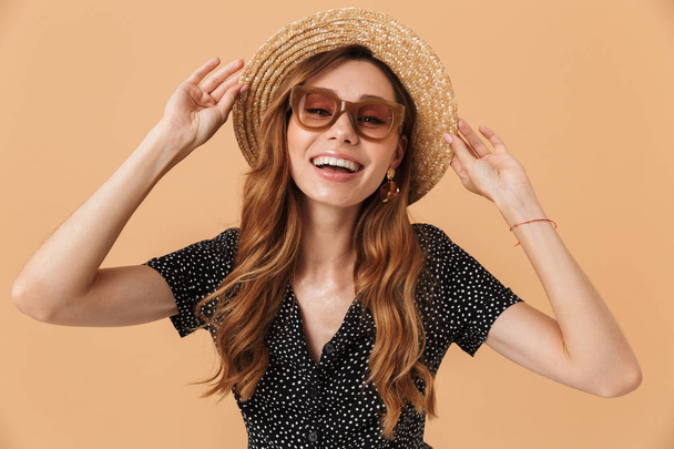 Photo of elegant european woman wearing sunglasses touching beautiful straw hat on head isolated over beige background - Foto, Imagen