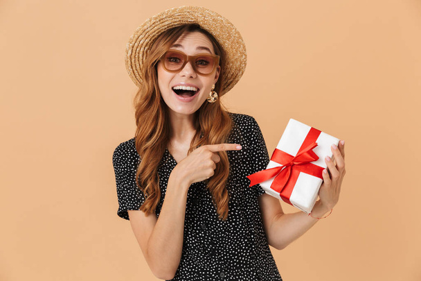 European summer woman 20s wearing straw hat and sunglasses smiling while holding present box with red ribbon isolated over beige background - Foto, Imagen