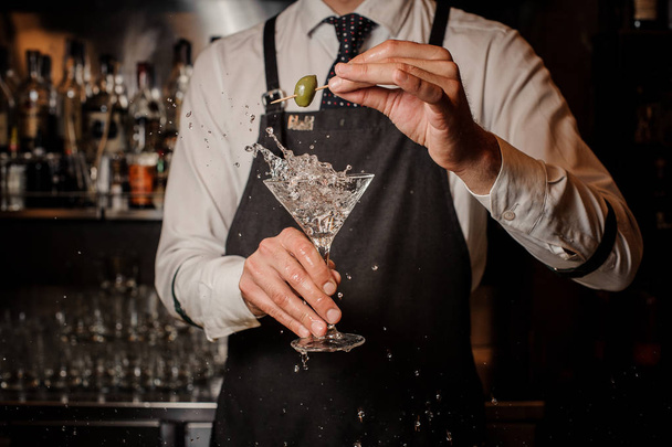 Bartender holding a splashing fresh and strong summer martini cocktail with a green olive against the bar - Фото, изображение