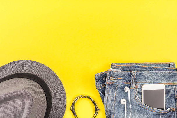 Flat lay of blue jeans, grey hat and smartphone headphones on bold yellow paper background with copy space. Overhead view of woman casual outfit. Trendy hipster look top view - Zdjęcie, obraz