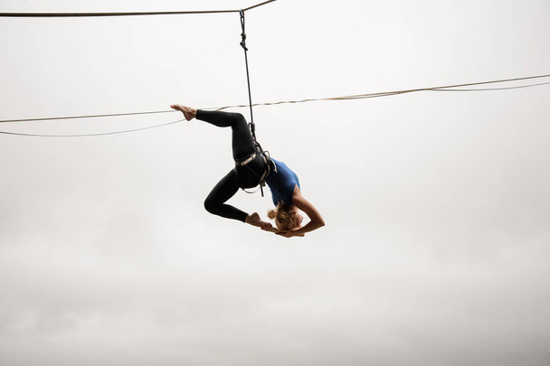Beautiful blonde girl balancing high on a slackline against the grey sky and town on rainy summer day - Фото, изображение