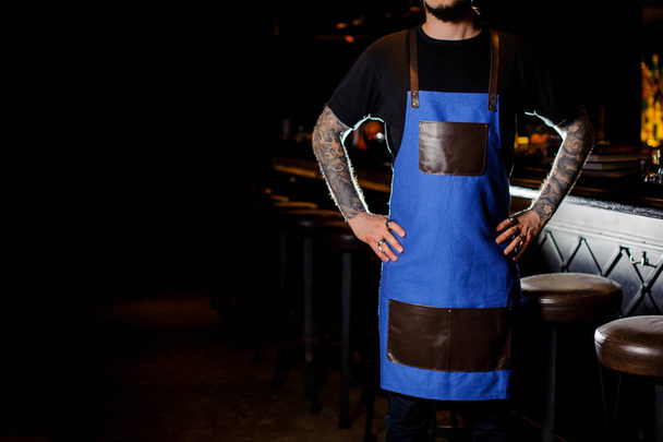 Barman with tattoo dressed in blue and brown apron standing near the dark bar counter in night club - Photo, Image