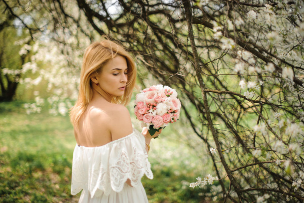 Back view of a young blonde woman in a white dress with bouquet near the blooming cherry tree - Fotó, kép