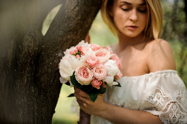 Portrait of a young blonde woman in a white dress with a bouquet of flowers standing near the tree - Fotoğraf, Görsel
