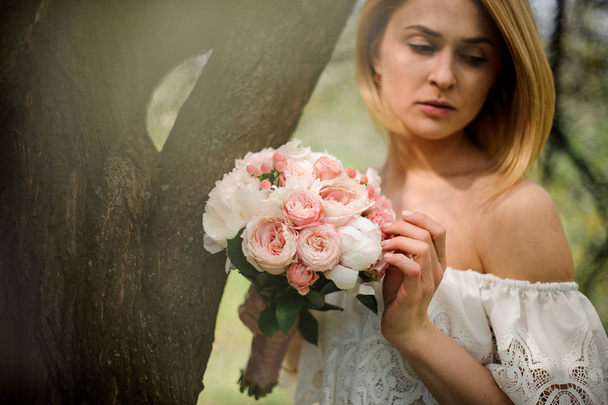 Tender portrait of a young blonde woman in a white dress with a bouquet of flowers standing near the tree - Fotó, kép