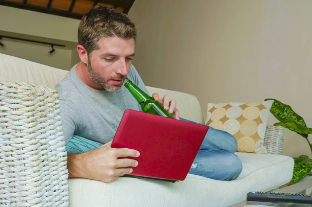 lifestyle indoors portrait of young attractive and handsome happy man lying at home sofa couch working online with laptop computer using netbook relaxed comfortable drinking beer bottle  - Foto, imagen