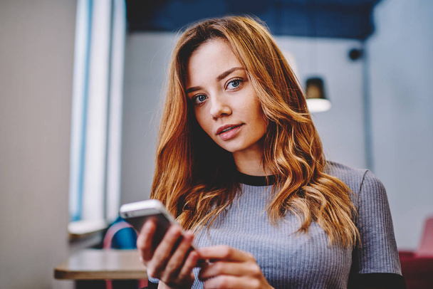 Close up portrait of beautiful millennial hipster girl looking at camera while waiting telephone call indoors, young attractive female student using mobile phone for chatting sitting at campus - Foto, afbeelding