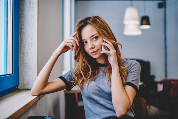 Portrait of beautiful hipster girl using 4g connection for communicated with friend via cellphone sitting at campus, charming teenage woman looking at camera and making telephone conversation indoors - Фото, изображение
