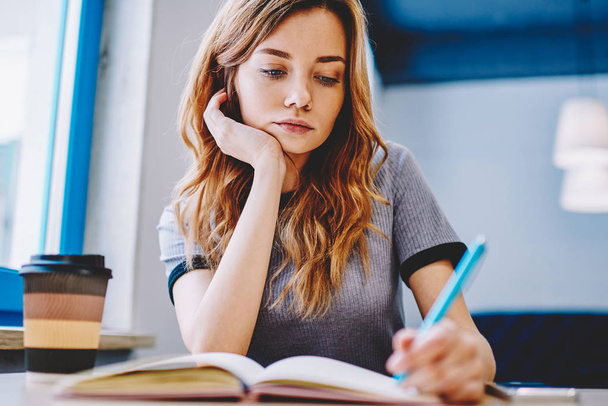 Young clever tired female student thinking and writing school homework in textbook for education, thoughtful woman sitting at desktop and spending time for course work indoors notes main theses - Fotó, kép