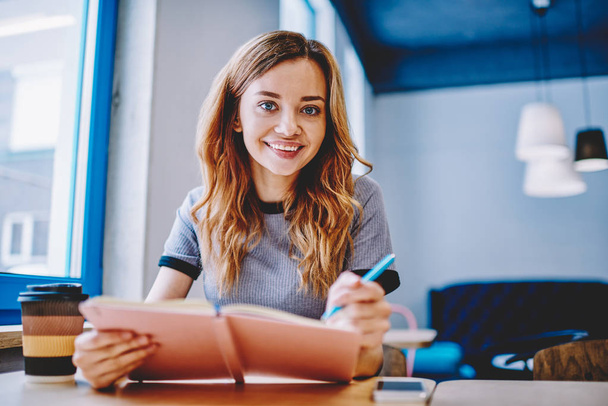 Portrait of positive female student sitting at table in coffee shop and enjoying autodidact,successful hipster girl making research for project in university and looking at camera in break of studying - Фото, изображение