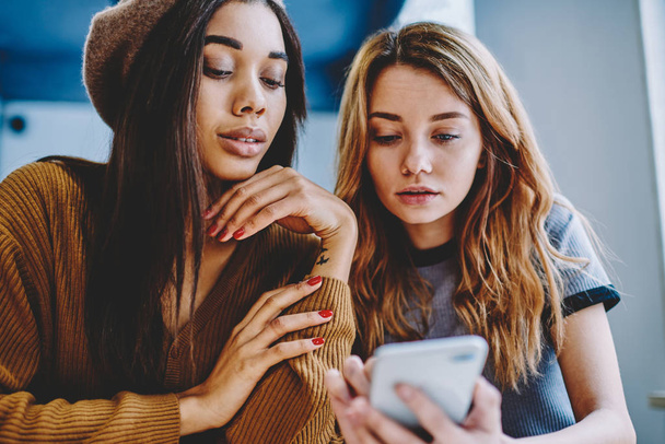 Serious female friends reading text message from friend using application for chatting on smartphone device, two concentrated millennial hipster girls searching information via cellular phone - Foto, afbeelding