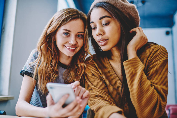 Portrait of happy successful hipster girl holding smartphone gadget in hand and sitting near female friend, positive smiling women searching information via multimedia application on cellular - Foto, immagini