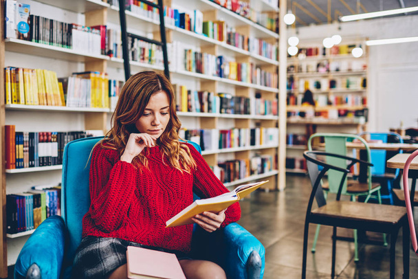 Young attractive caucasian hipster girl reading interesting best seller pondering on information sitting in comfortable chair with bookshelves on background, concept of knowledge and education - 写真・画像