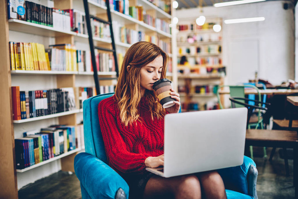 Smart skilled female copywriter drinking morning takeaway coffee sitting at chair with laptop device with bookshelves on background, clever hipster girl enjoying caffeine beverage after e learning - Foto, Imagen