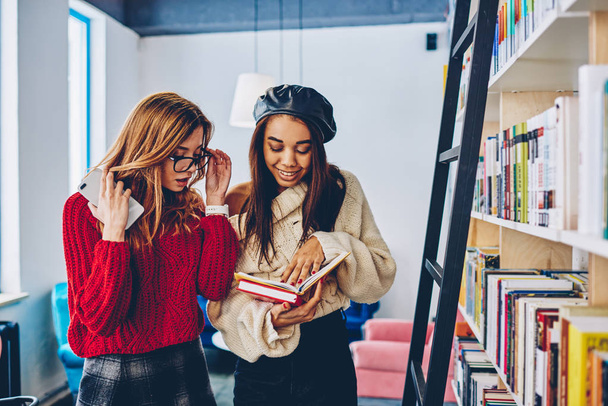 Happy clever female friends searching information on literature enjoying education in modern bookstore, positive trendy hipster girls reading novel standing near bookshelves in academy library - Фото, изображение
