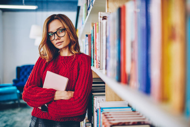 Puoli pituus muotokuva houkutteleva hipster tyttö silmälasit visio korjaus tilalla oppikirja koulutukseen ja katsot kameraa, nuori valkoihoinen viehättävä nainen viettää aikaa kirjakaupassa
 - Valokuva, kuva