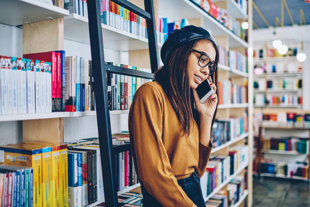 Smiling young hipster girl standing near bookshelves and enjoying friendly cellular call at bookstore, successful african woman making positive telephone conversation using 4g connection indoors - Photo, image
