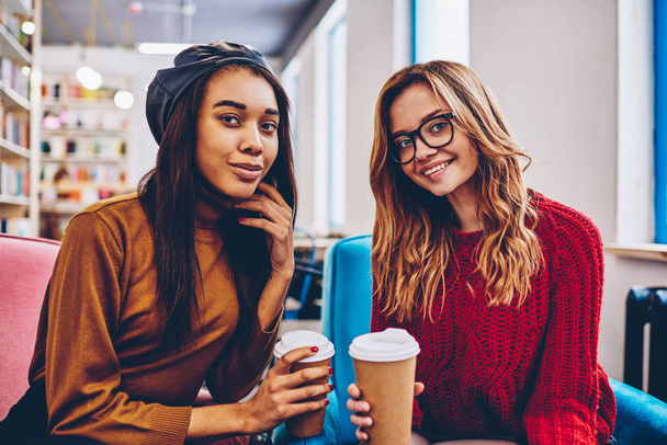 Portrait of successful multicultural hipster girls looking at camera and enjoying coffee break indoors, happy smiling female teenagers in trendy apparel holding takeaway cup at campus on leisure - Foto, Imagem