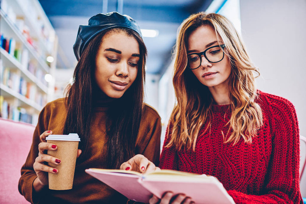 Young hipster girls reading interesting literature together during coffee break indoors, african woman with caffeine beverage searching information in textbook sitting near caucasian friend in eyewear - Photo, Image