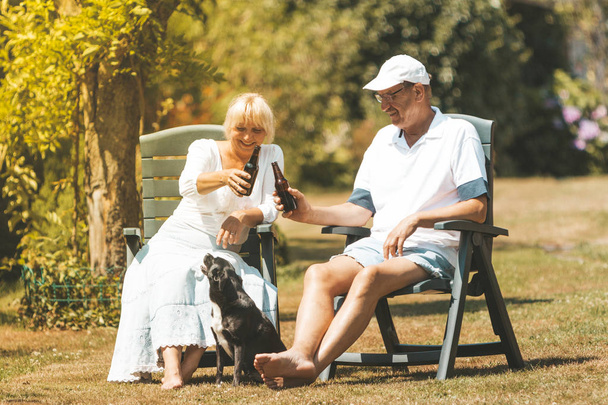 A retired couple is relaxing and having fun in the garden, drinking beer and playing with small dog - Φωτογραφία, εικόνα