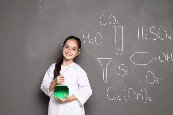 Little school child in laboratory uniform with flask of liquid and chemical formulas on grey background - Photo, image