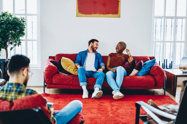Happy male and female multiracial friends having fun in modern designed apartment communicating in good mood, cheerful woman and man laughing during conversation on leisure resting on red sof - Zdjęcie, obraz