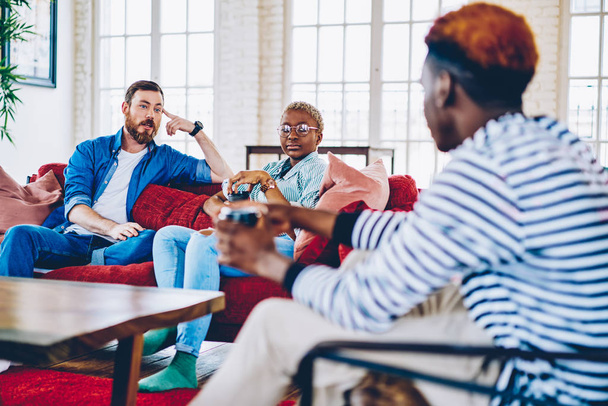 Group of male and female friends spending free time together at modern designed living room in apartment communicating with each other, multiracial hipsters having conversation at home interior - Photo, image