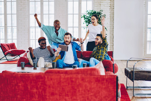 Overjoyed multiracial group of male and female fans cheering for soccer game watching on digital tablet together in loft interior, happy emotional hipsters expressing surprise with victory of contes - Fotoğraf, Görsel