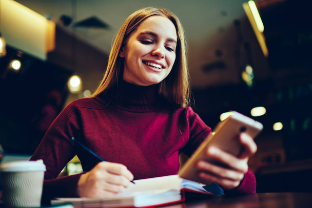 Happy cheerful woman making research for course work while received positive text message from friend via smartphone gadget, smiling female student using internet on cellular for planning trip - Фото, зображення